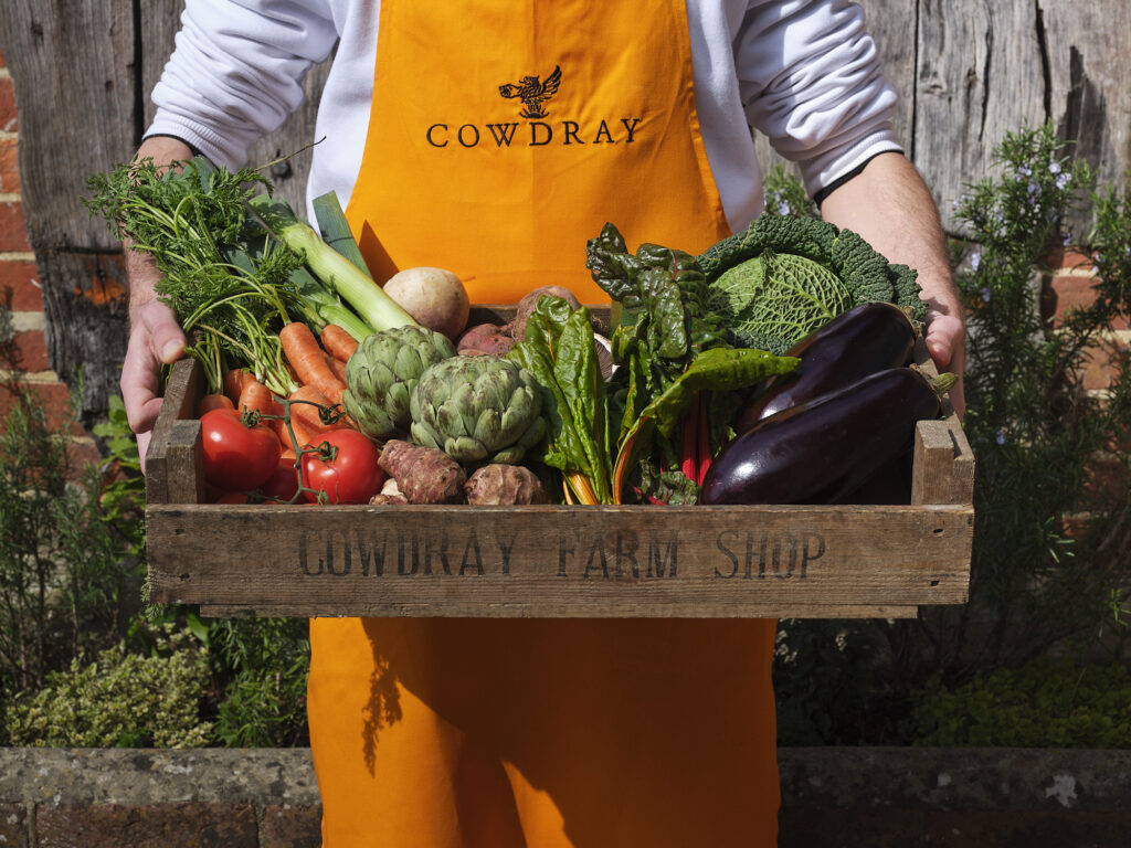 A closeup of a Cowdray Farm Shop staff member wearing a yellow Cowdray apron holding a wooden box full of fresh seasonal vegetables in Midhurst, West Sussex.