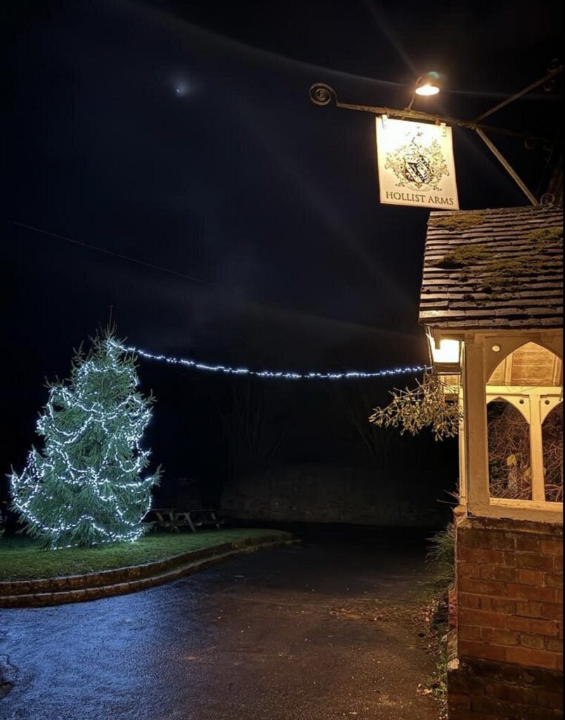 An exterior view of The Hollist Arms pub at night, showing a lit Christmas tree.