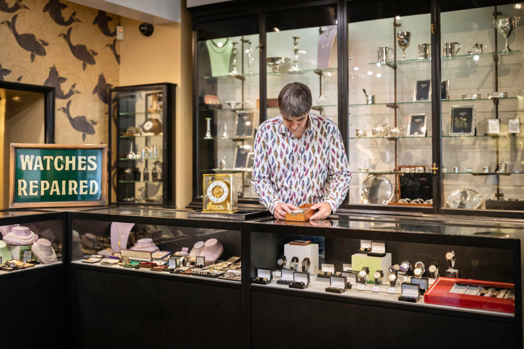 Inside J E Allnutt & Son Ltd shopfront in West Street, Midhurst with a shopkeeper behind the counter which shows vintage watches, jewellery and silver.