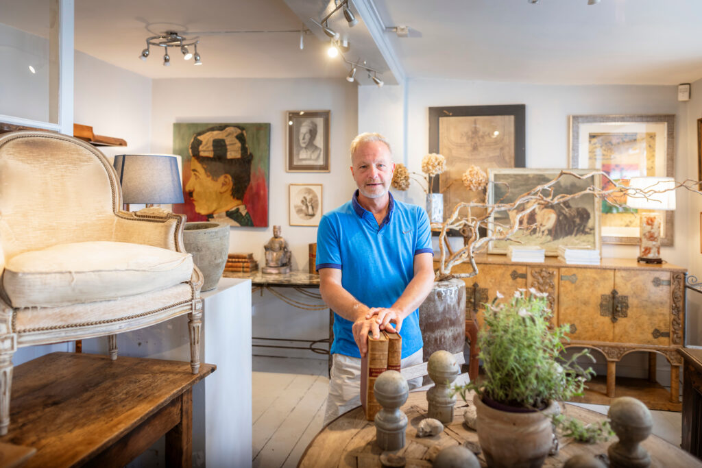 Inside of Olive Interiors in Midhurst, West Sussex with the shopkeeper surrounded by country house style furniture and antiques.