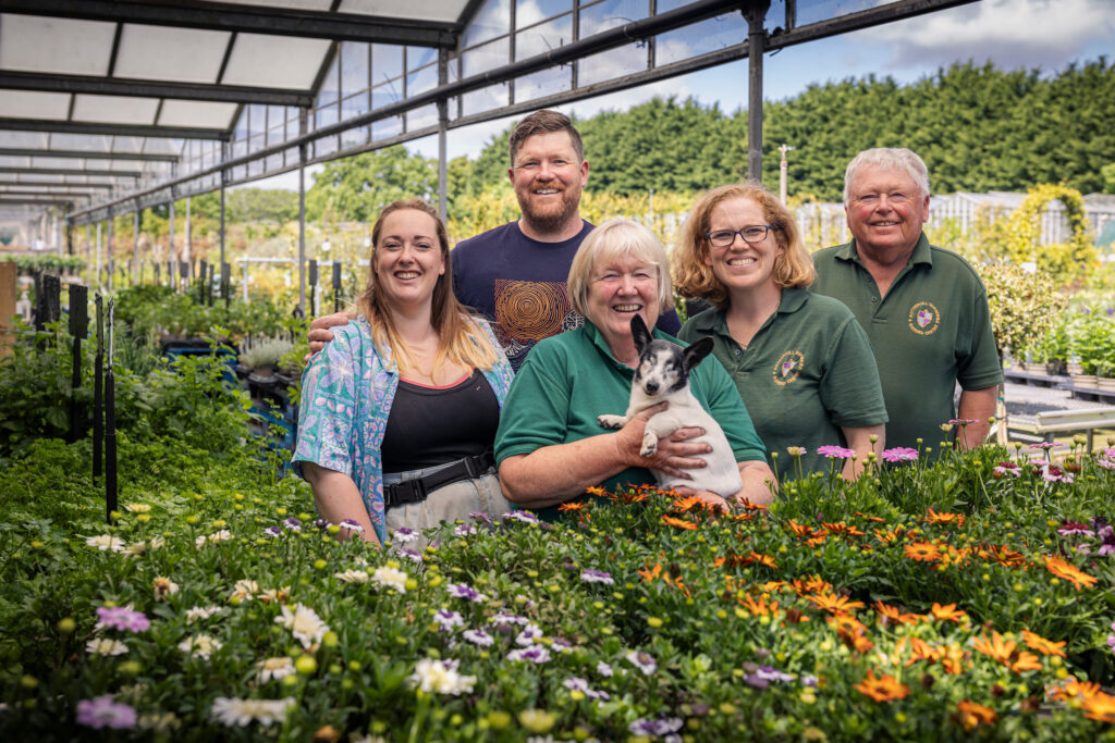 Five happy staff members of Rotherhill Nurseries and Garden Centre and a dog surrounded by growing flowers.