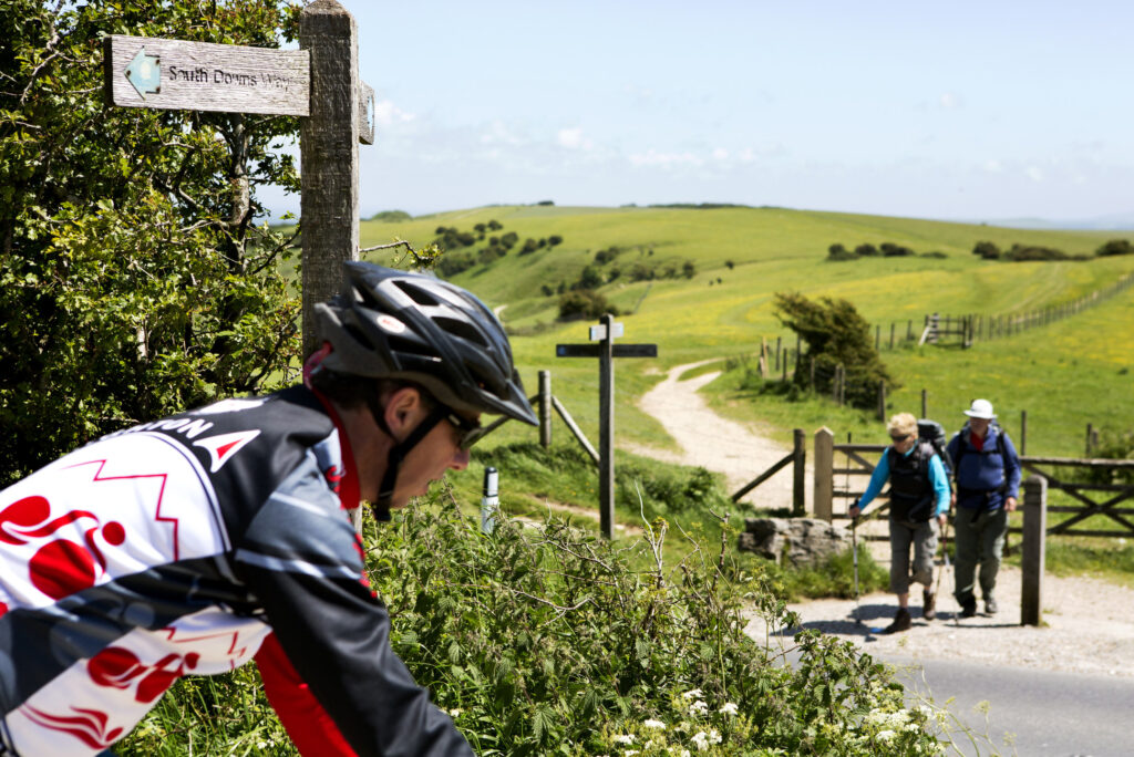 A cyclist cycling the South Downs Way with rolling green hills in the background with an elderly couple walking.
