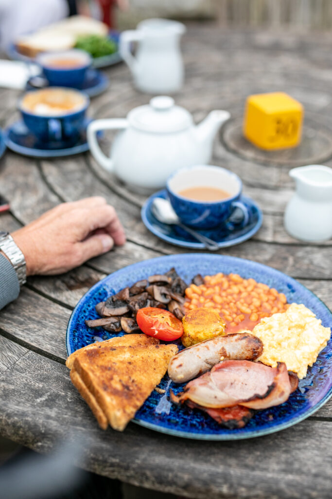 Closeup of an English breakfast and cups of tea made by Cowdray Farm Shop & Cafe, on an outdoor wooden table.